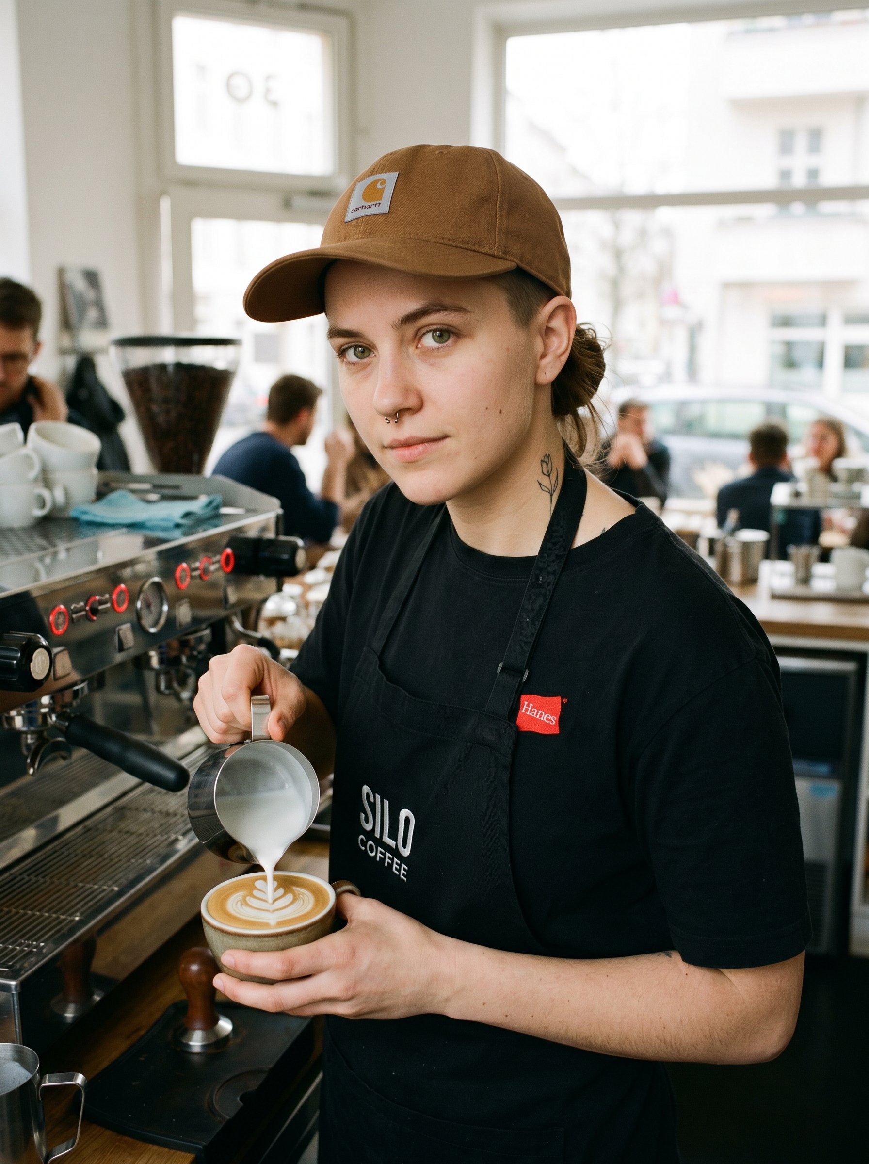 Closeup-portrait three-quarter same subject as portrait-tight, soft Saturday-Nachmittag-Café-Innen-light at the Silo-Coffee Boxhagener-Eck bar-station, she stands behind the LaMarzocco-Linea-PB-Espresso-Machine with her right hand mid-pour on a milk-pitcher creating a Tulip-Pattern Latte-Art-Linie plus her left hand stabilising the espresso-cup, the brown Carhartt-WIP-Cap visible plus the small fine-line spine-tattoo just visible at the back of the neck where the T-shirt collar pulls down (Latte-Art-Linien-Design), eye-contact direct