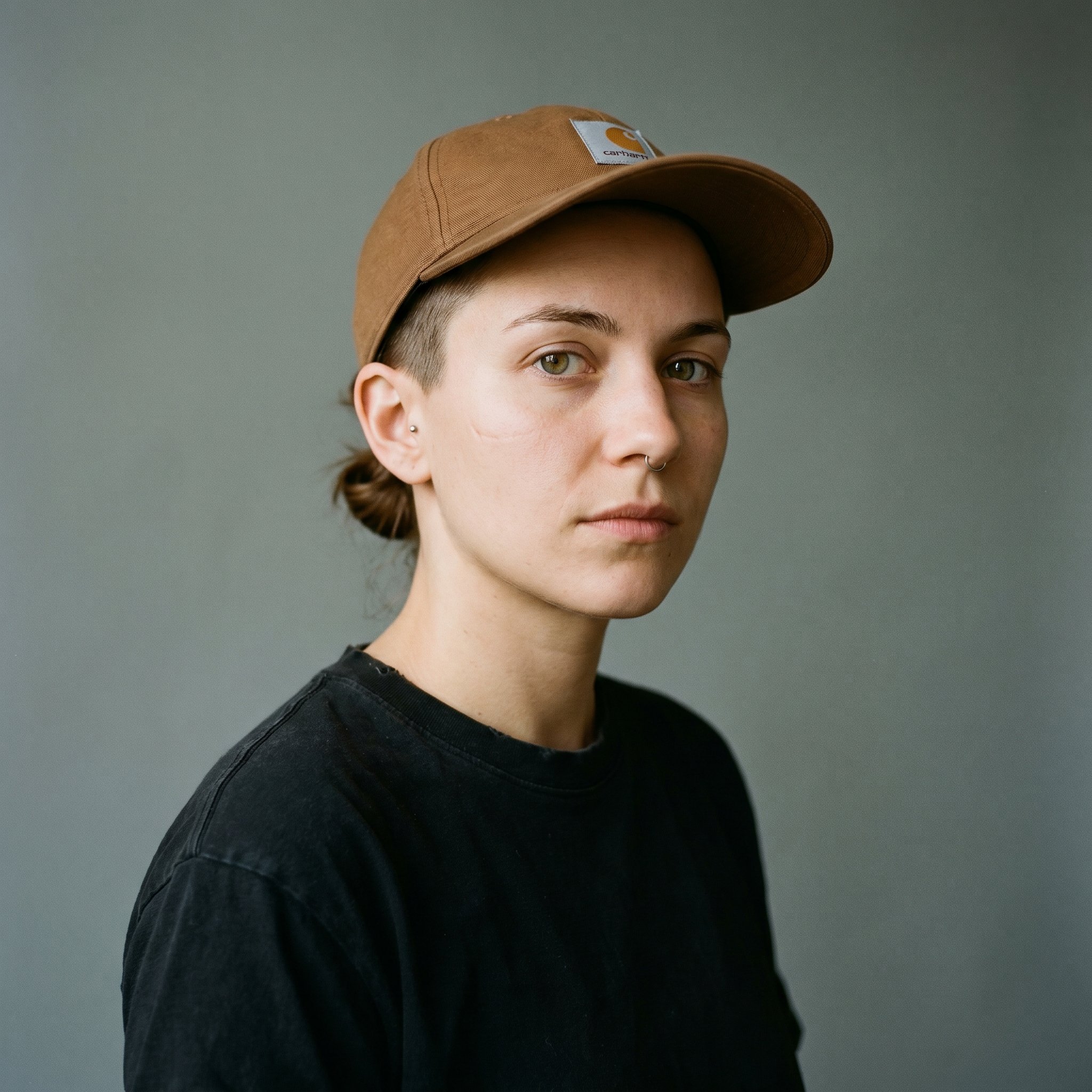 Portrait-Tight Frame, 1:1 frontal, neutral background, Nina Adam 25 Berlin-Friedrichshain Silo-Coffee-Barista Boxhagener-Eck Specialty-Coffee Q-Grader 2024, calm direct eye contact, brünette undercut hair longer on top swept-back, Carhartt-WIP-Cap brown the Café-Werk-Uniform-Cap, plain black washed-soft cotton T worn three years
