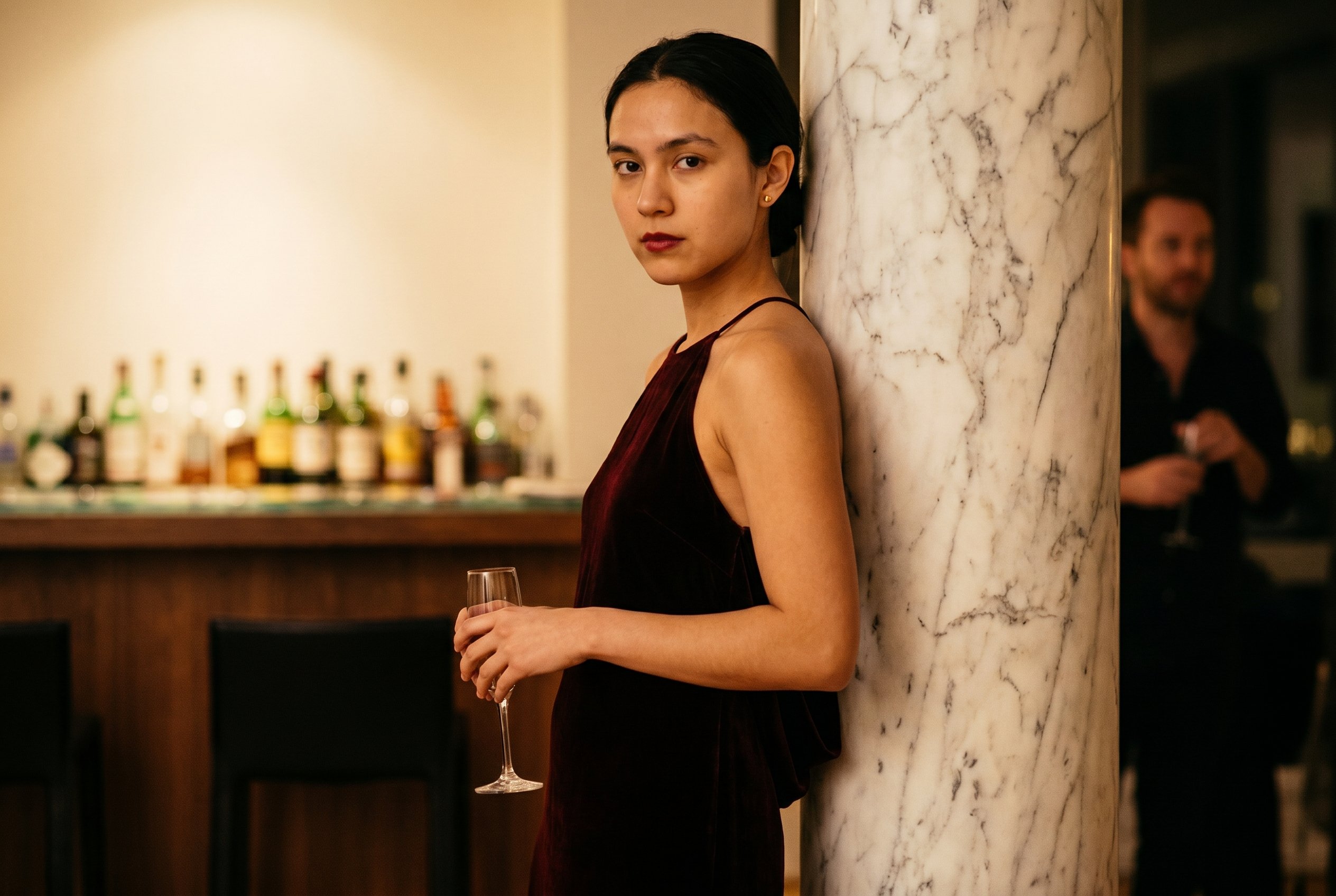 Tribeca penthouse interior, leaning against a marble column near the bar with a champagne-flute lowered to the hip, conversation off-frame