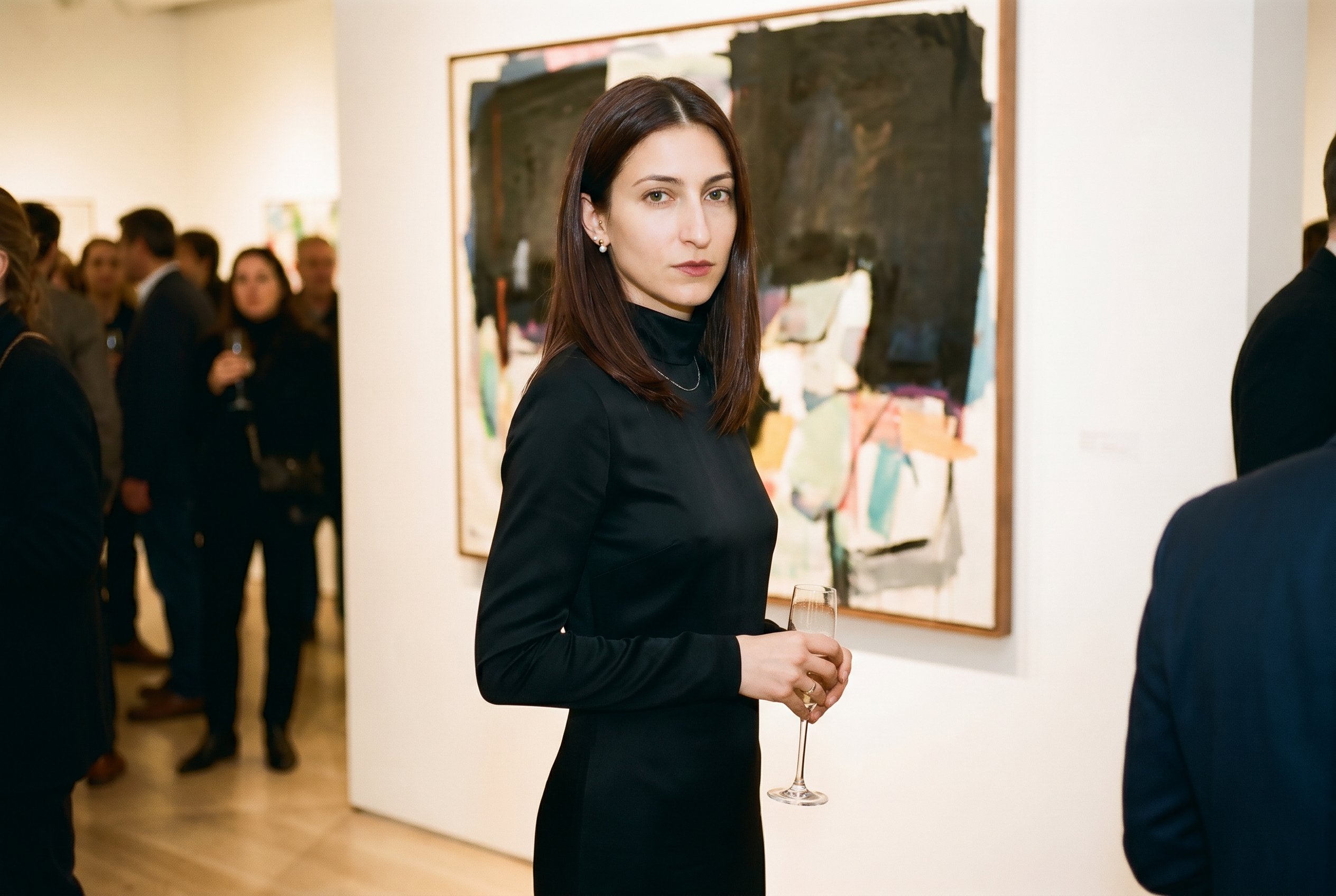 Bowery-and-Stanton gallery main-room at a Thursday-night vernissage, standing in front of a large-format painting with a flute of champagne in the right hand, half-turned toward the wall and half-toward the camera