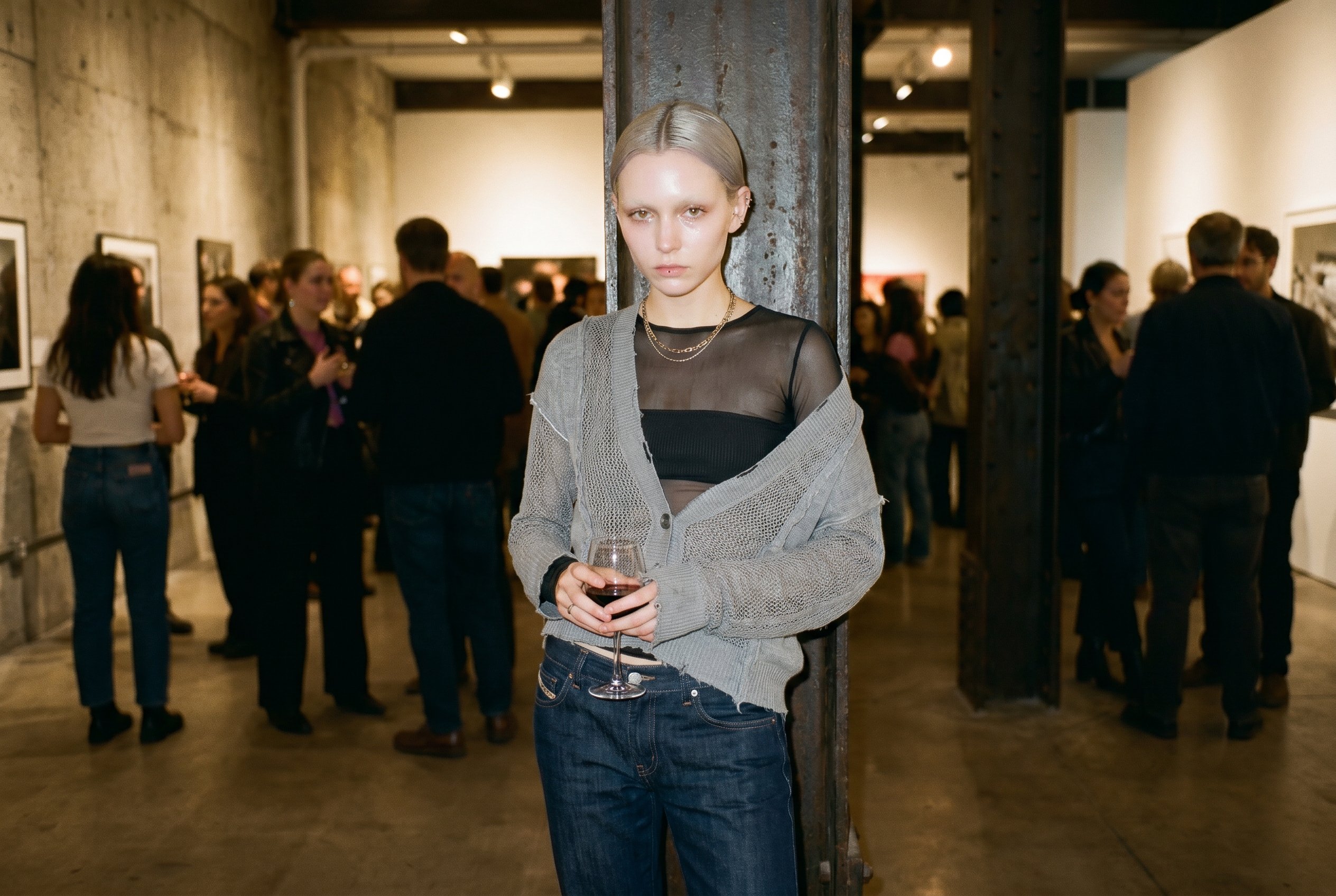 Meatpacking District Washington-Street storefront gallery interior, planted mid-room near a raw exposed steel I-beam, weight on the back foot, wine glass held low at hip-height, the Eckhaus-Latta-mesh-and-knit-layer visible at the shoulder, looking past the camera with neither smile nor scowl, the warm gallery-crowd in soft tungsten bokeh behind