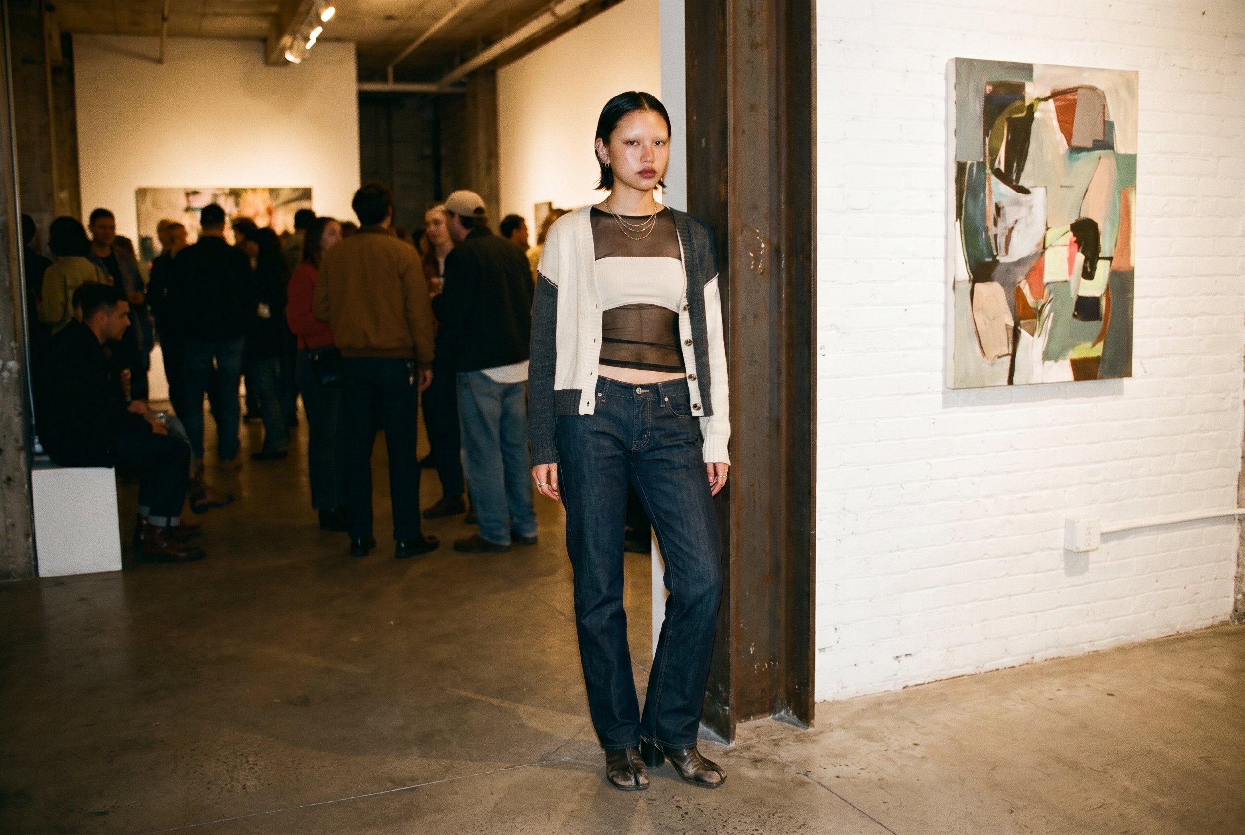 Meatpacking District Washington-Street storefront gallery interior, planted centred at the far end of the room near a raw exposed steel I-beam beside one of the artist's mid-size paintings, weight on the back foot, the Eckhaus-Latta-mesh-and-knit-layer visible at the shoulder, looking past the camera with neither smile nor scowl, raw-concrete floor and white-painted brick in soft fall-off