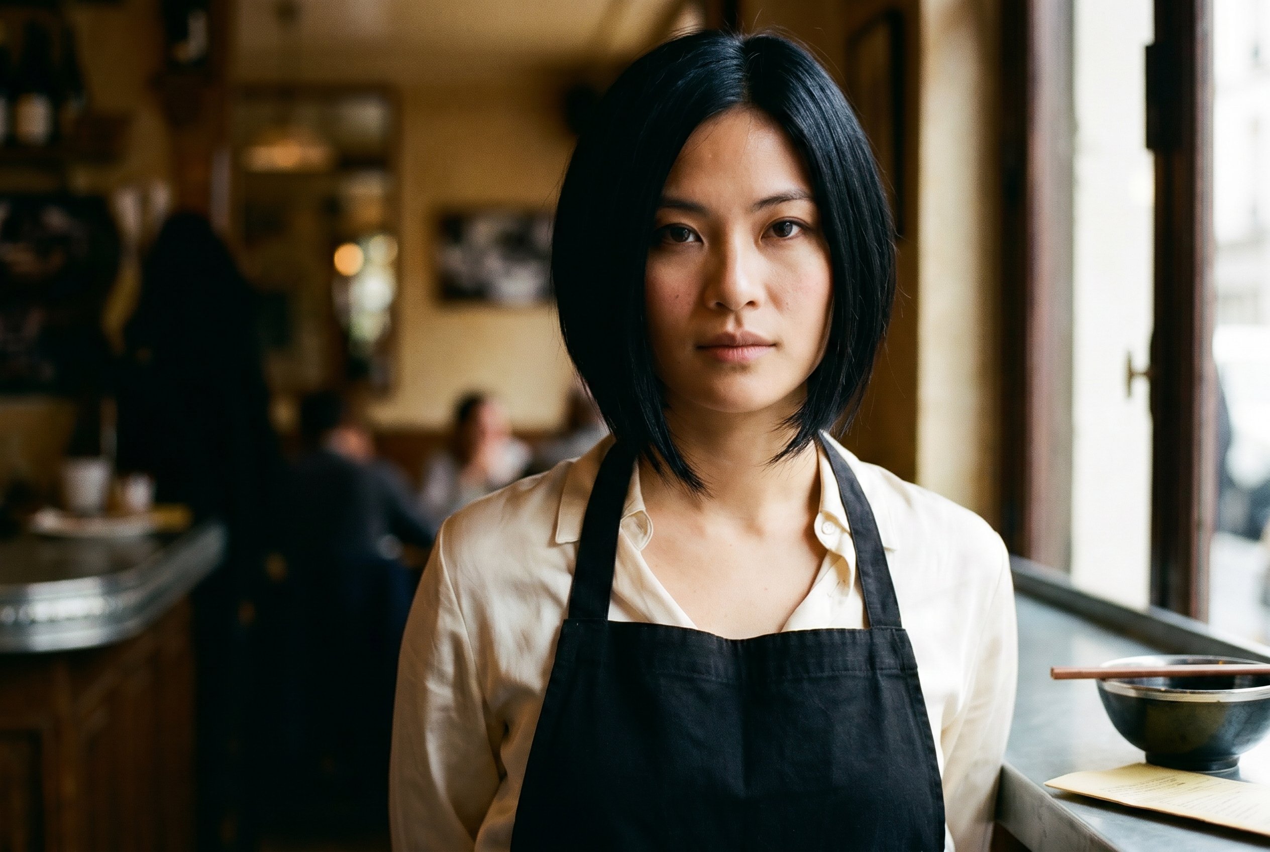 Closeup — Linh at the counter, the warm soft-focus bistro-interior in the background, the small lacquered black ceramic phở-bowl with a hand-cut bamboo chopstick at the right edge, her grandfather's small folded square of yellowed 1968-Hanoi recipe-paper tucked under the bowl-rim