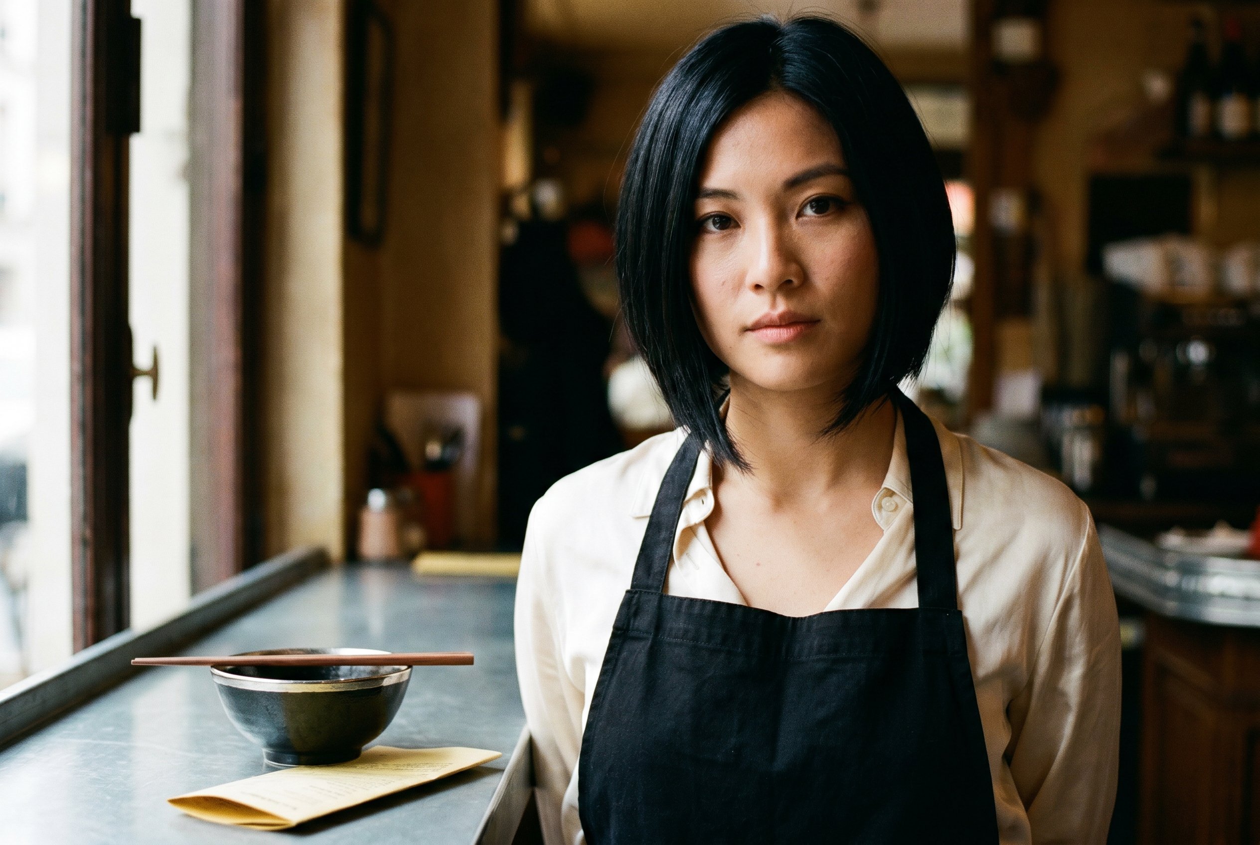 Portrait — Linh at the two-seat zinc-counter of her newly-opened Chinatown-13e phở-bistro, the Subject-Lock anchor for the wider context-frames below