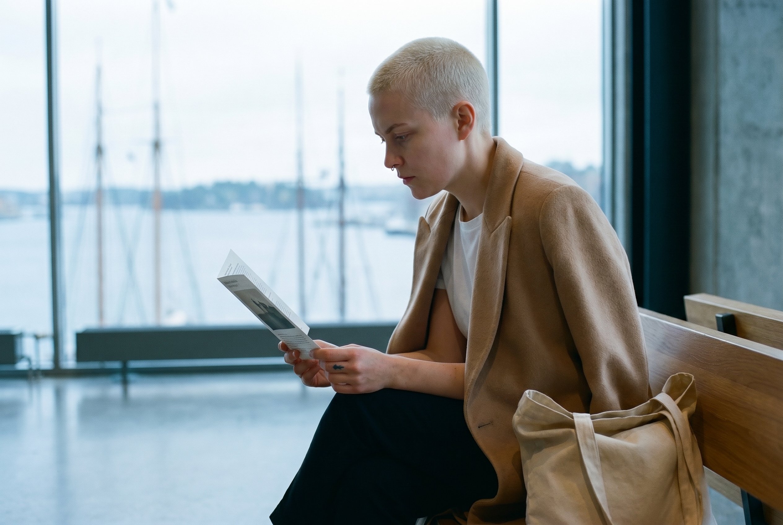 Closeup — Maja on the second-floor foyer bench at Fotografiska, reading the Atoosa Farahmand show-pamphlet, the Studio-Brus canvas tote at her side, the harbor-light cool-blue from the window behind