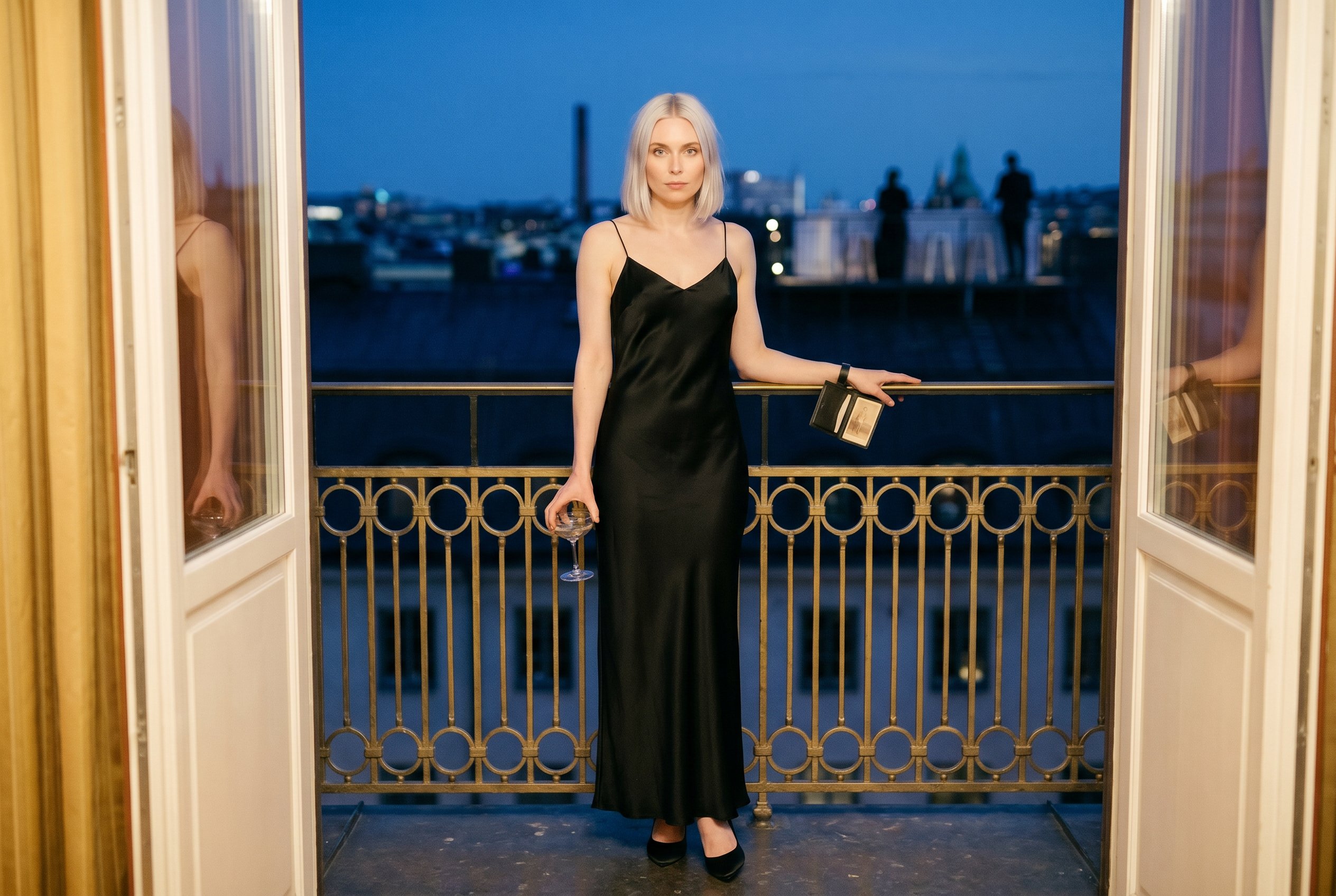 Context — Astrid at full body on the Berns Salonger Roof-Terrace with the Stockholm Berzelii-Park and the Strandvägen-Nybroviken-night-skyline in deep soft focus behind, the late-evening Stockholm-May-cool-blue rooftop-light, the gala-amber-light bleeding from the rooftop-terrace-doors at camera-front-left